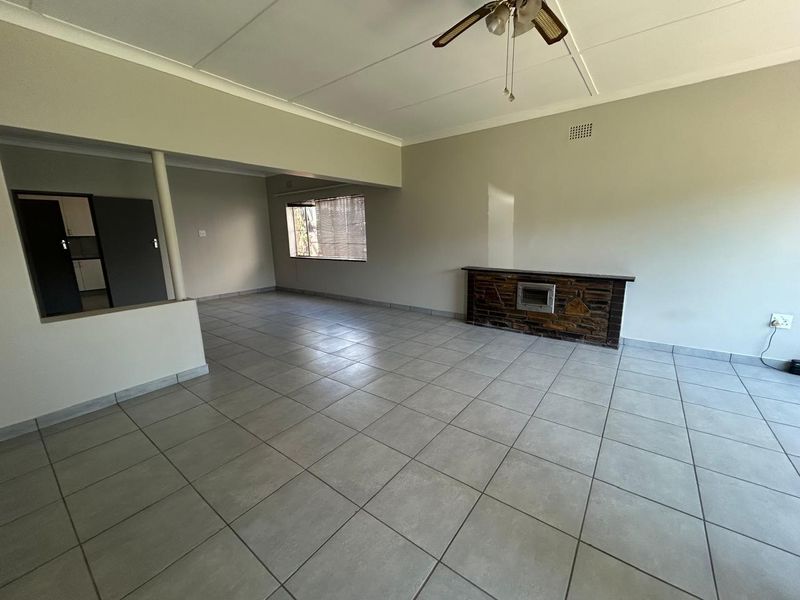 Spacious open-plan living area featuring neutral tiled flooring, a stone-clad fireplace, and a ceiling fan, offering a versatile layout for modern living.