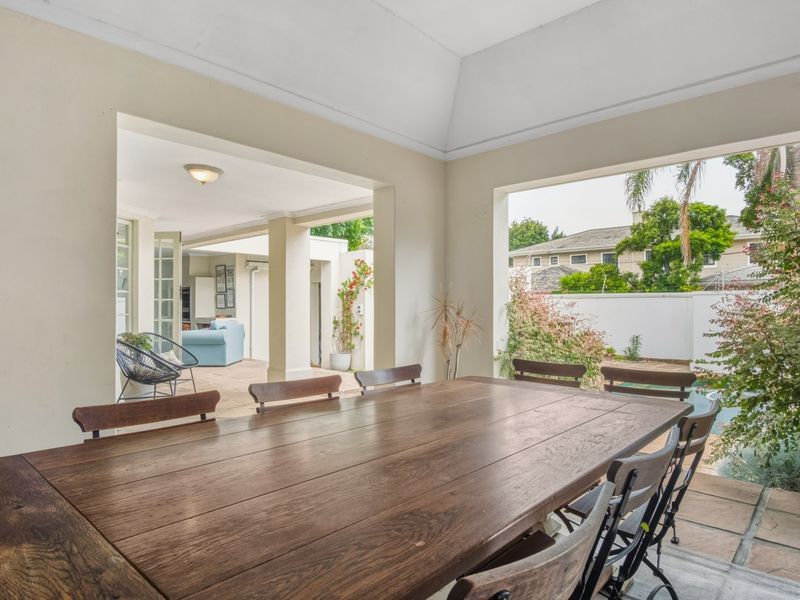 A spacious covered patio featuring a vaulted ceiling, stone flooring, and structural columns, offering a seamless transition to the outdoor garden and pool area.