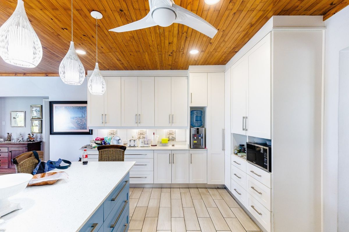 Modern kitchen featuring white cabinetry, stone countertops, a central island, and a distinctive timber-paneled ceiling with integrated lighting and a ceiling fan.