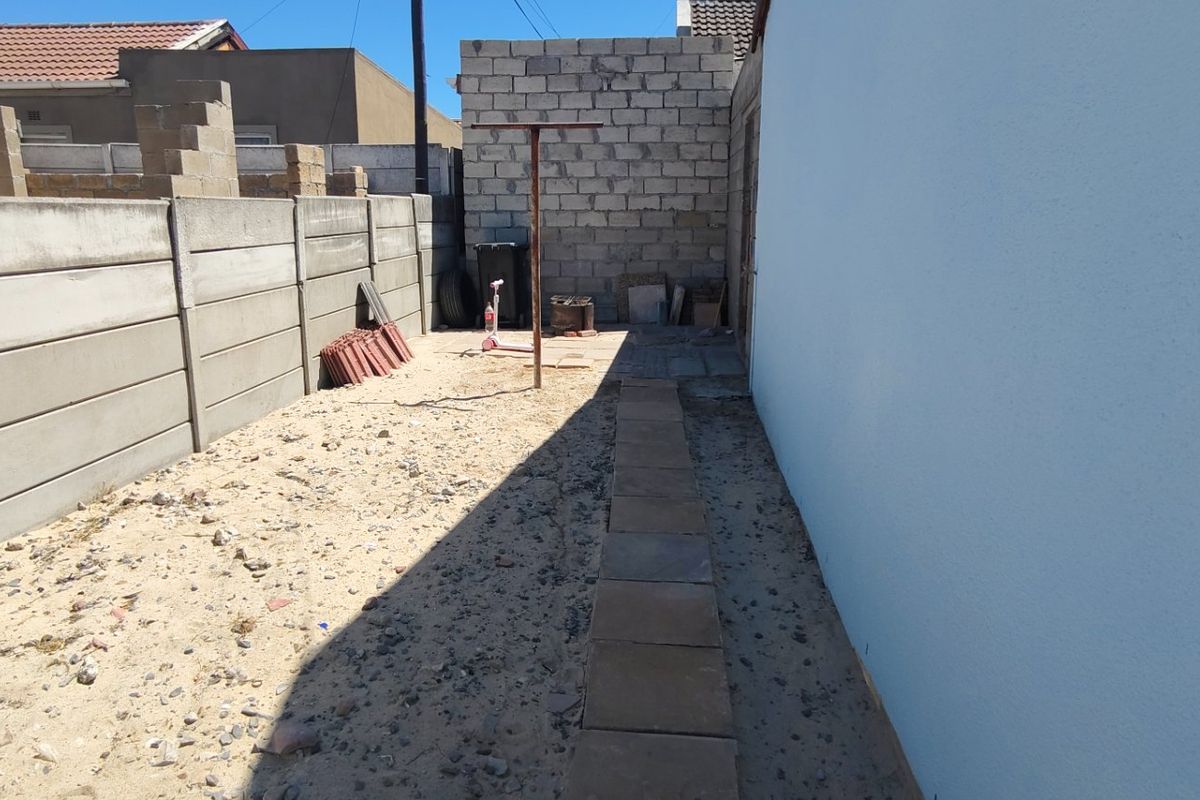 A rear exterior area featuring a precast concrete boundary wall, an unfinished concrete block wall, and a simple paved walkway leading through a sandy yard.