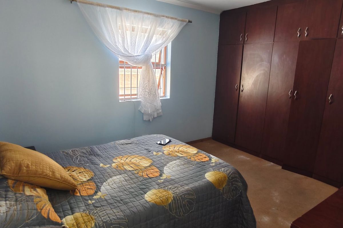 A bedroom featuring built-in wooden cupboards and a window with security bars, offering a functional space with potential for cosmetic updates.