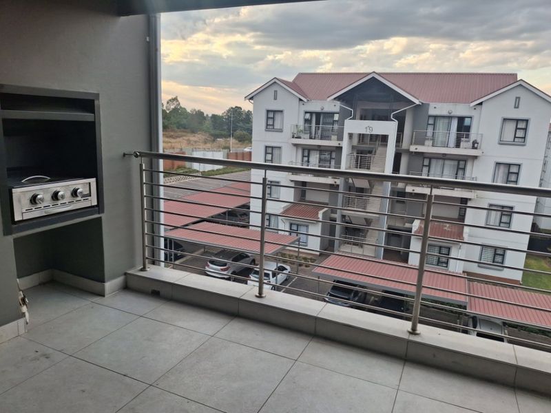 Covered balcony featuring a built-in braai, tiled flooring, and a stainless steel balustrade, overlooking the apartment complex parking area.