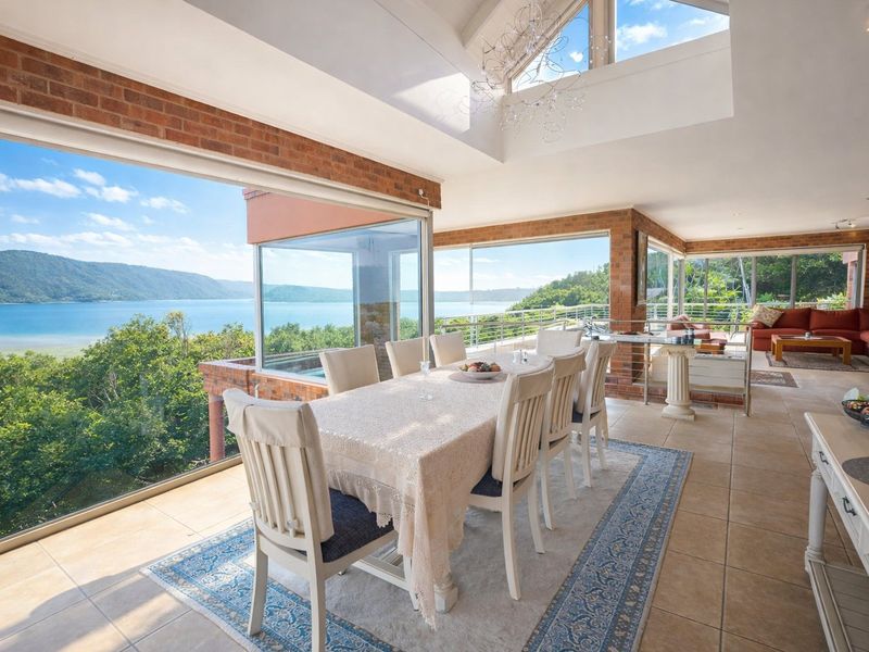 Spacious open-plan dining and living area featuring vaulted ceilings, exposed brickwork, and expansive glass walls offering panoramic water views.