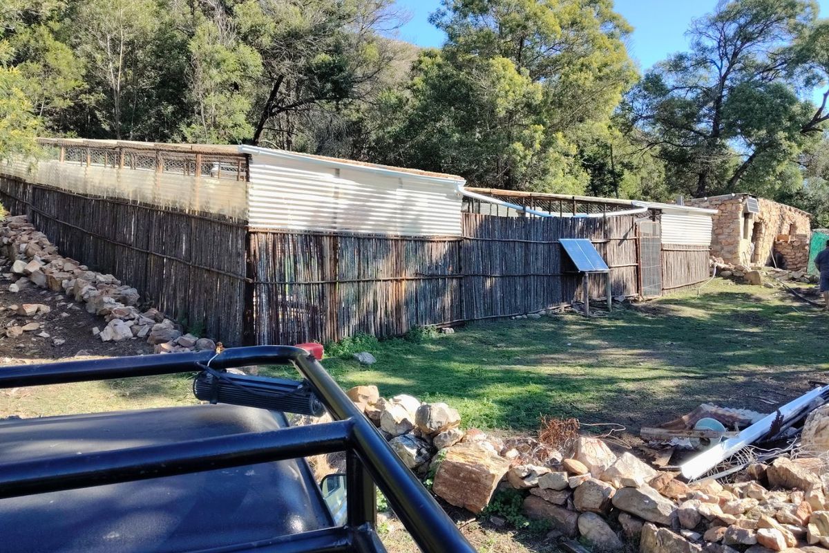 Exterior view of a farm structure featuring corrugated iron roofing, reed fencing, and a solar panel installation set against a natural landscape.