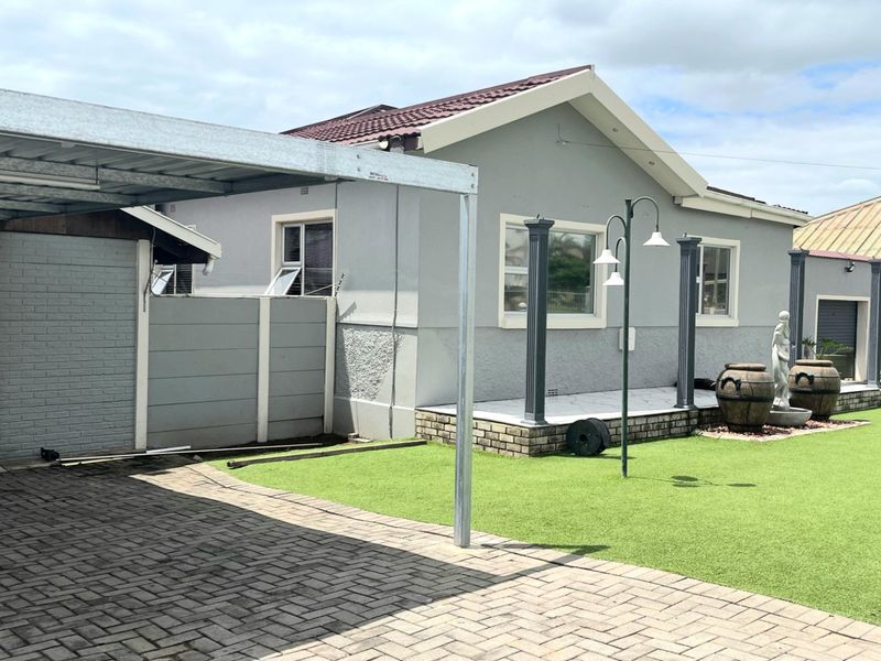 Rear exterior view featuring a paved driveway, carport, artificial lawn, and a tiled patio area with decorative pillars.