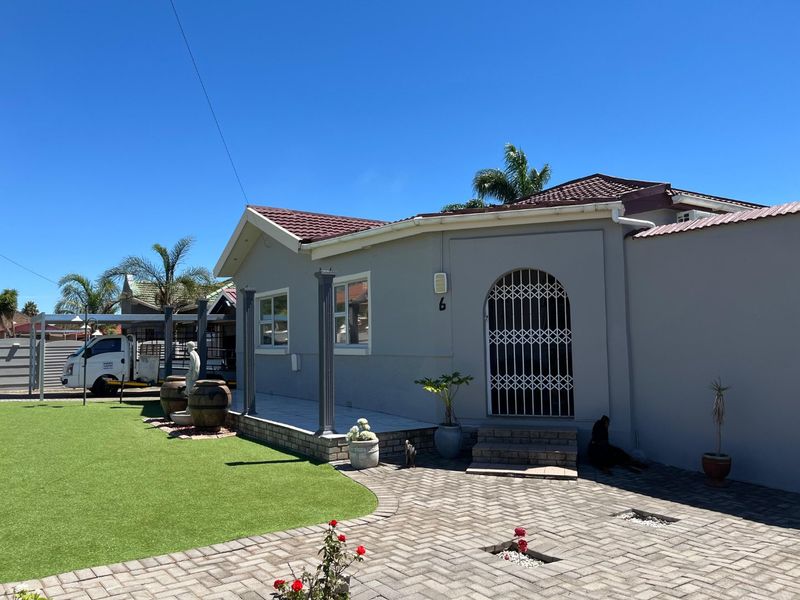 Front exterior view of a single-story house featuring a paved driveway, artificial lawn, covered patio area, and secure gated entrance.