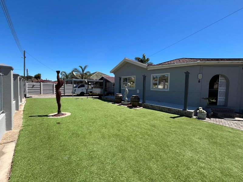 A well-maintained front exterior view featuring a neat artificial lawn, paved driveway, secure perimeter walling, and a covered carport area.