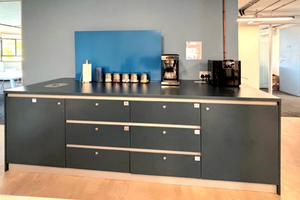 A functional office kitchenette area featuring dark cabinetry, a blue splashback, and integrated counter space for beverage preparation.
