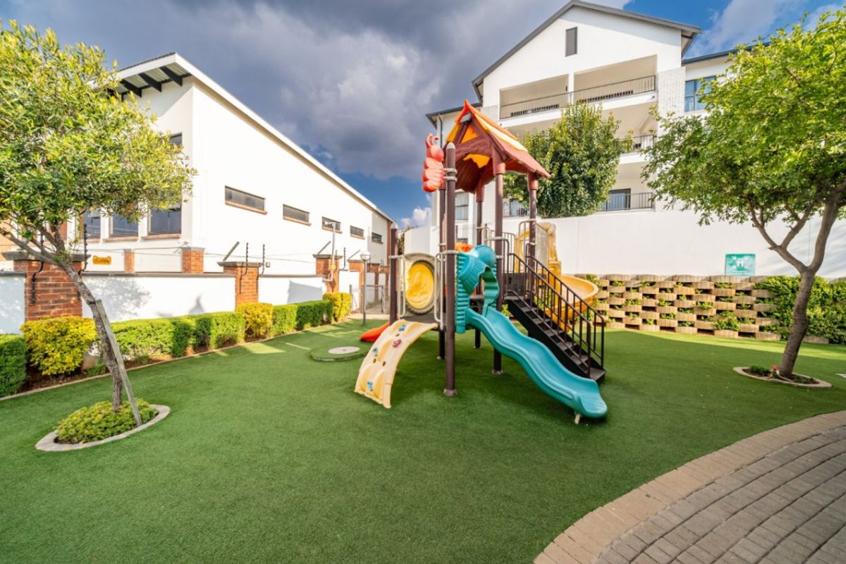 A well-maintained communal garden area featuring artificial turf, a children's playground structure, and secure perimeter walling within an apartment complex.