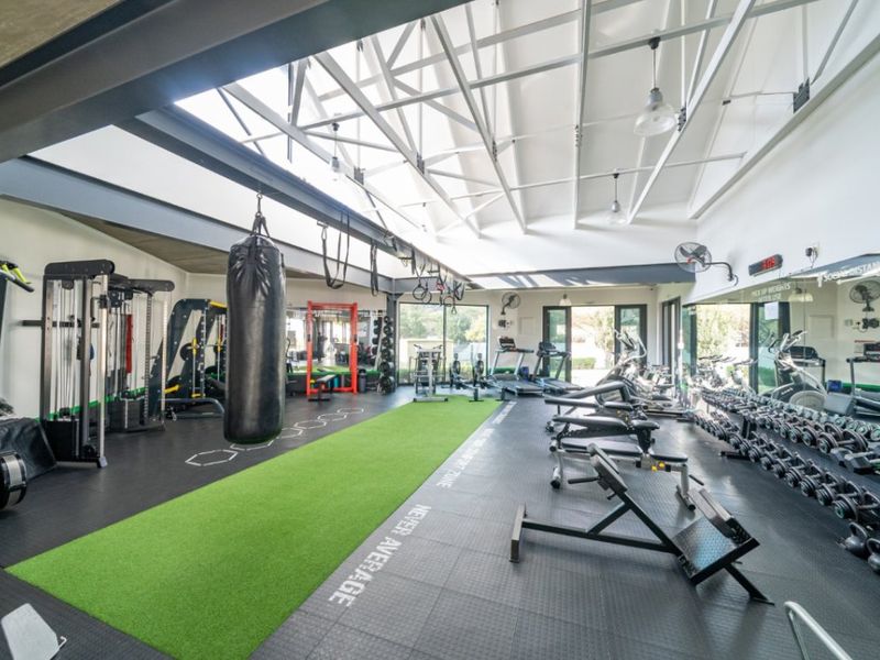 A well-equipped, modern gym featuring exposed steel roof trusses, high ceilings, rubberized flooring, and full-length mirrors, providing a professional fitness environment.