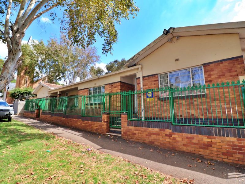 Front exterior view of a brick house featuring secure perimeter fencing, security bars on windows, and a paved sidewalk approach.