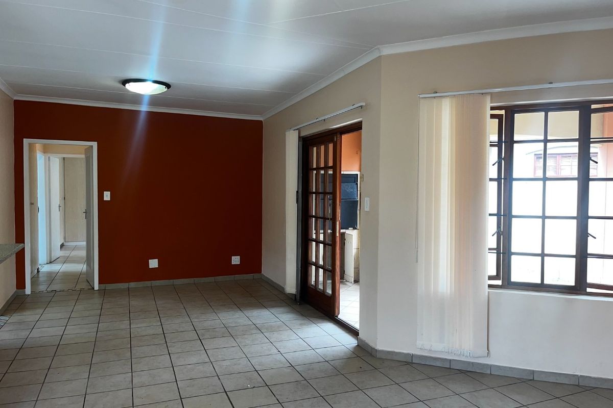 An empty living room featuring tiled flooring, a wooden framed glass door leading to an adjacent space, and a large window with vertical blinds.