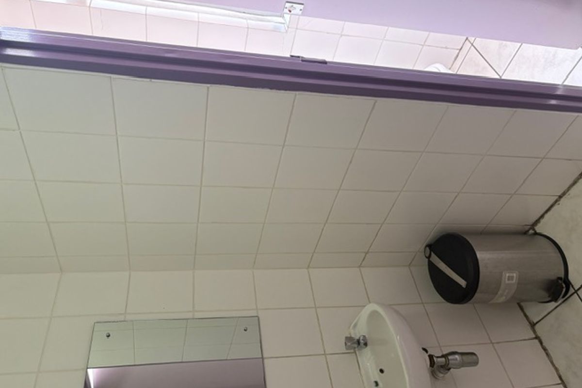 A compact bathroom featuring a wall-mounted basin, tiled walls and flooring, and a simple mirror, presenting a functional but dated commercial space.