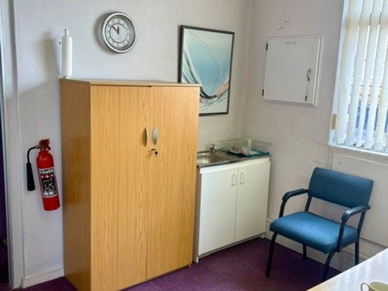 A small utility or office space featuring a built-in sink cabinet, drop ceiling, and vertical blinds, showing signs of wall plaster cracking.