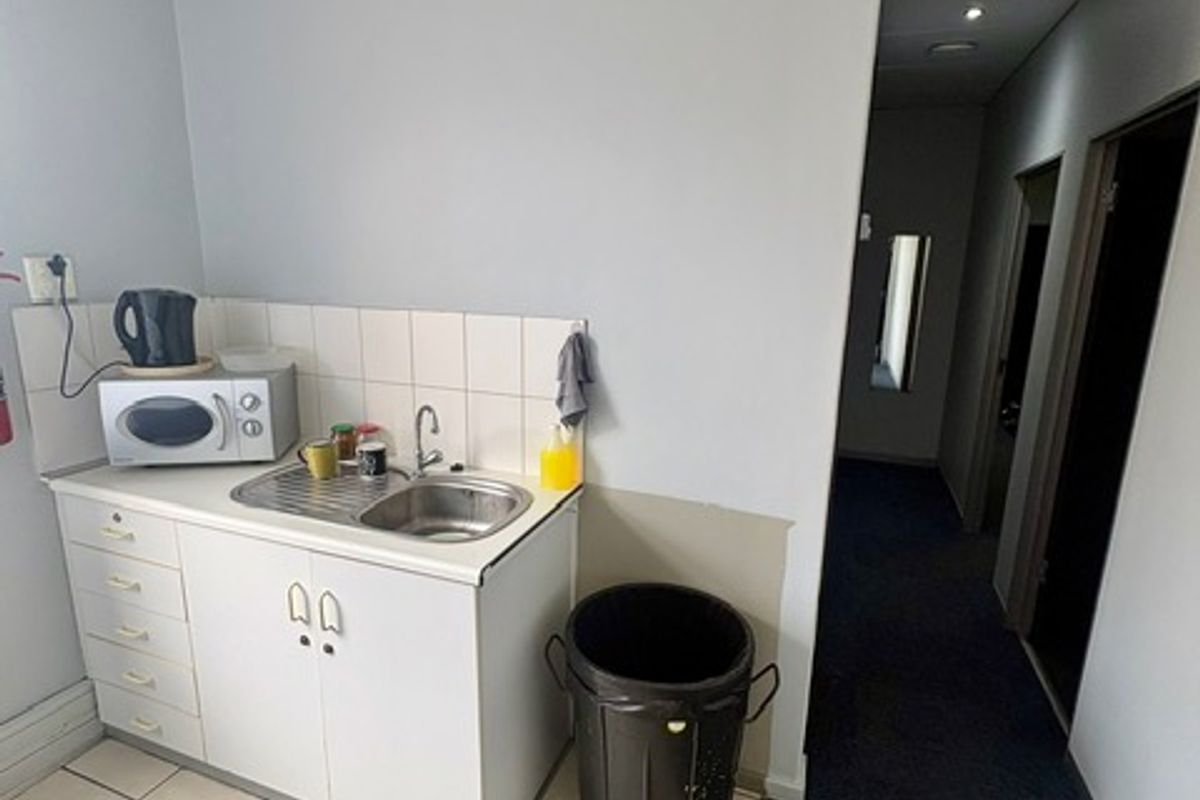 A compact commercial kitchenette featuring a stainless steel sink, tiled splashback, and built-in cabinetry, located adjacent to a carpeted hallway.