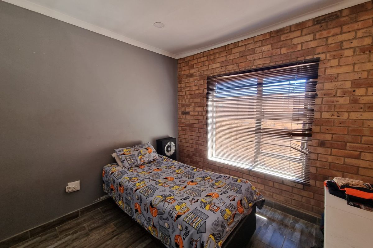 A bedroom featuring an exposed brick feature wall, tiled flooring, and a window fitted with venetian blinds.