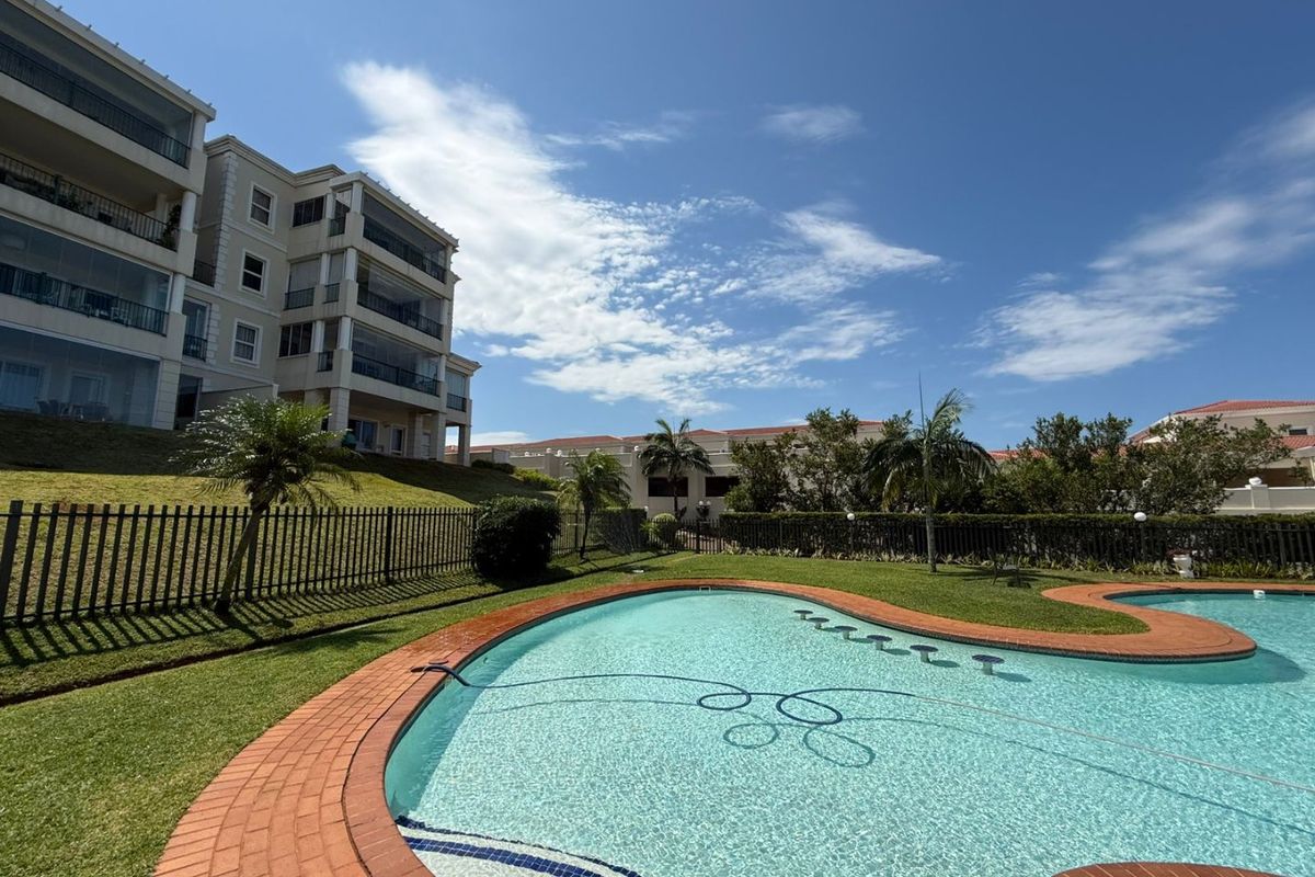 A well-maintained communal swimming pool area featuring brick coping, surrounded by landscaped lawns and a secure perimeter fence, set against the apartment building.