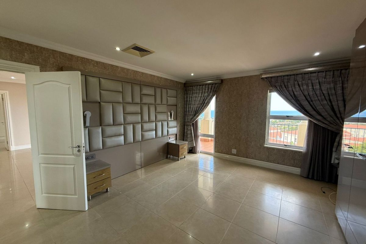 Spacious bedroom featuring a custom upholstered headboard wall, tiled flooring, recessed ceiling lighting, and large windows offering scenic coastal views.