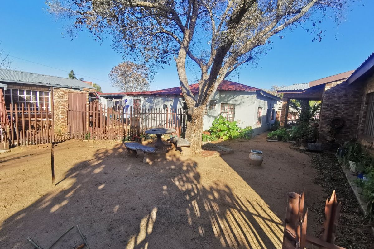 A spacious rear yard area featuring mature trees, brick and metal fencing, and a concrete patio space with potential for landscaping improvements.