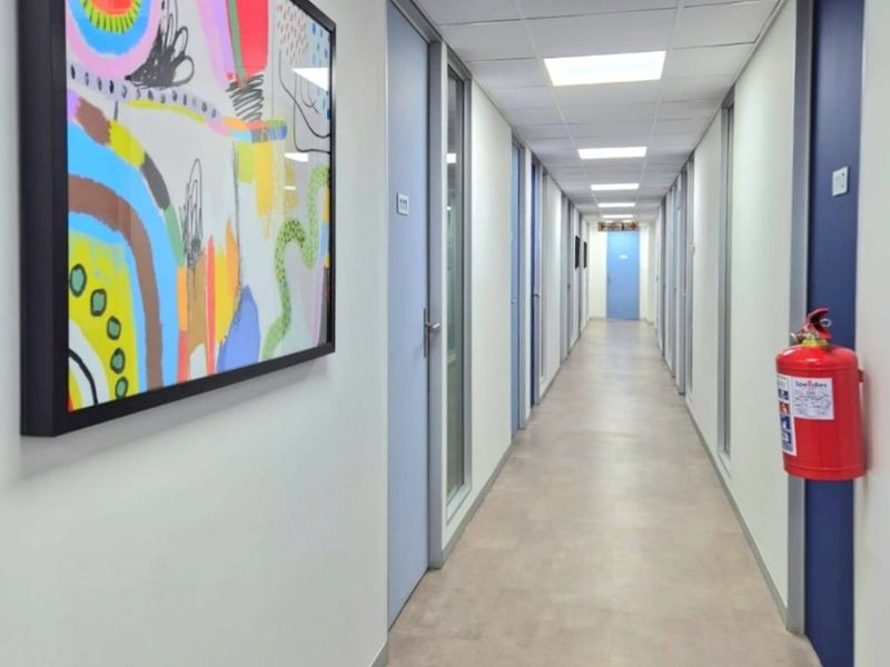 A clean, well-lit commercial office hallway featuring suspended ceiling tiles, recessed lighting, fire safety equipment, and multiple access doors.