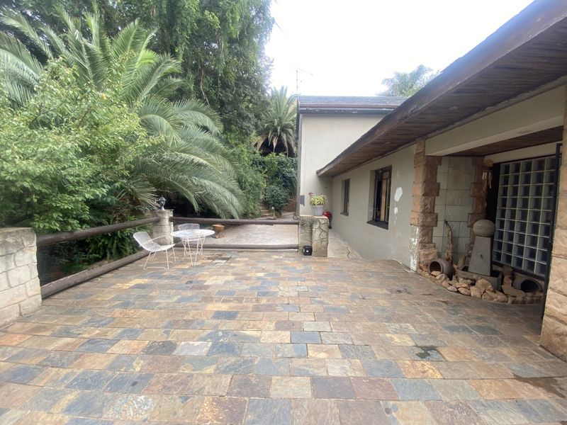 A spacious outdoor patio area featuring natural stone paving, stone pillar accents, and a covered walkway, set against a lush garden backdrop.