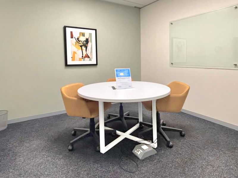 A functional office meeting room featuring carpeted flooring, a wall-mounted glass whiteboard, and neutral wall finishes.
