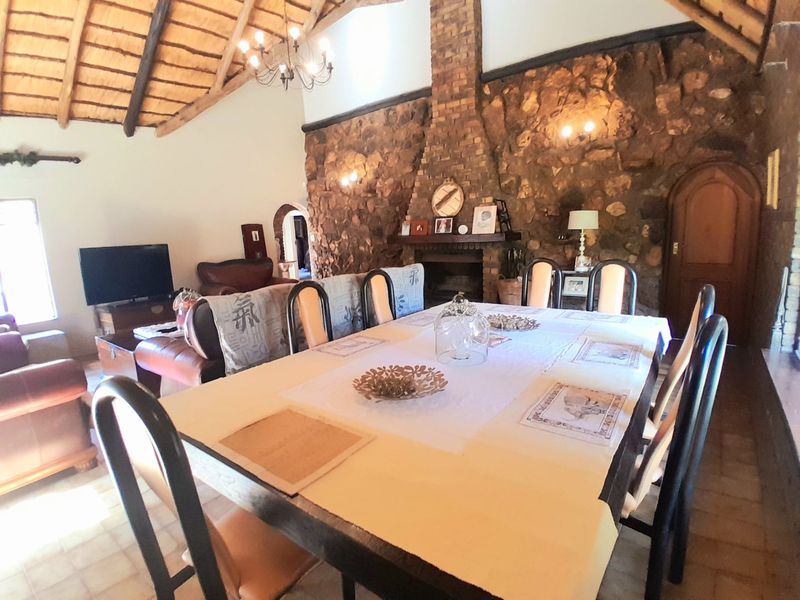 Spacious open-plan living and dining area featuring a high thatched ceiling with exposed rafters, a prominent stone feature wall, and a traditional brick fireplace.