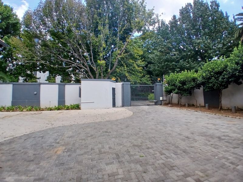 A secure front entrance featuring a paved driveway, perimeter wall with electric fencing, and a metal security gate surrounded by mature trees.