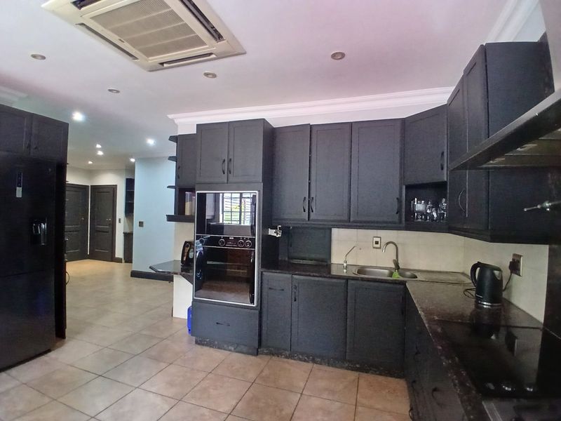 Modern kitchen featuring dark cabinetry, built-in oven, integrated hob, stainless steel sink, and ceiling-mounted air conditioning, set against tiled flooring.