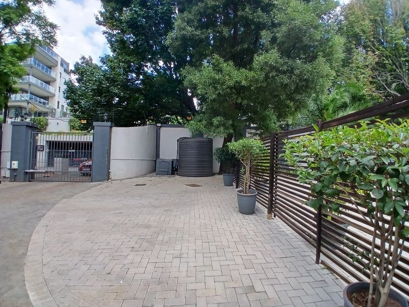 A paved front driveway area featuring a secure electric gate, perimeter walling, and a water storage tank, surrounded by established trees and fencing.