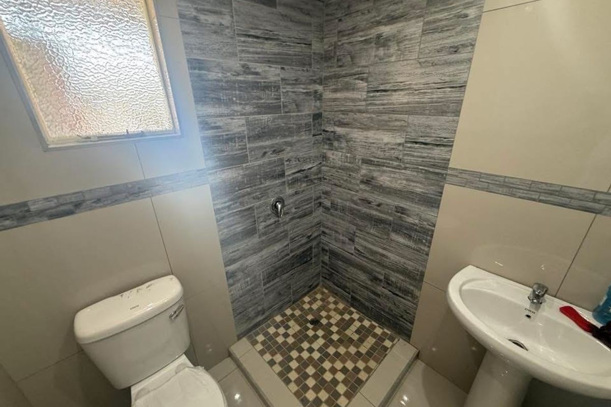 A clean, modern bathroom featuring a tiled shower enclosure, pedestal basin, and toilet with neutral wall tiles and a textured feature wall.