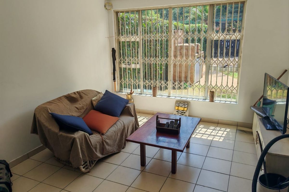 A living room featuring ceramic floor tiles and a large window fitted with security bars, providing natural light and garden views.
