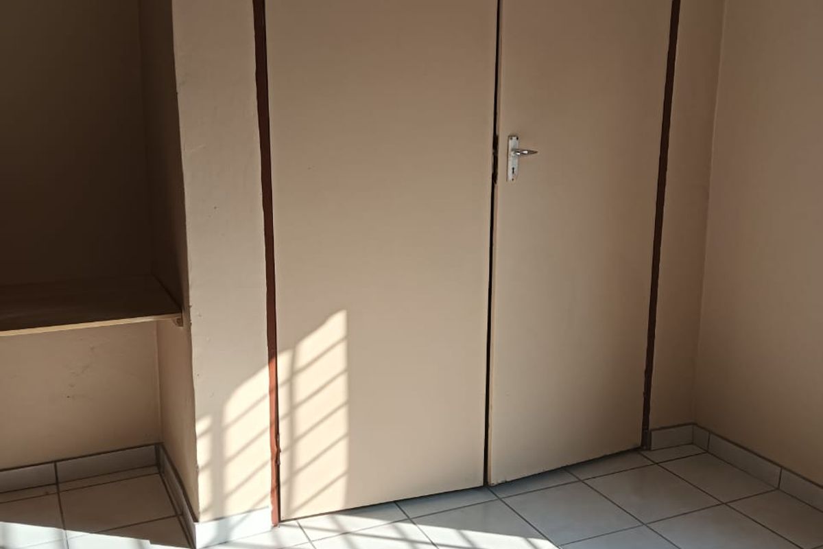 A bedroom featuring built-in cupboards with overhead storage and tiled flooring, illuminated by natural light through a window with security bars.