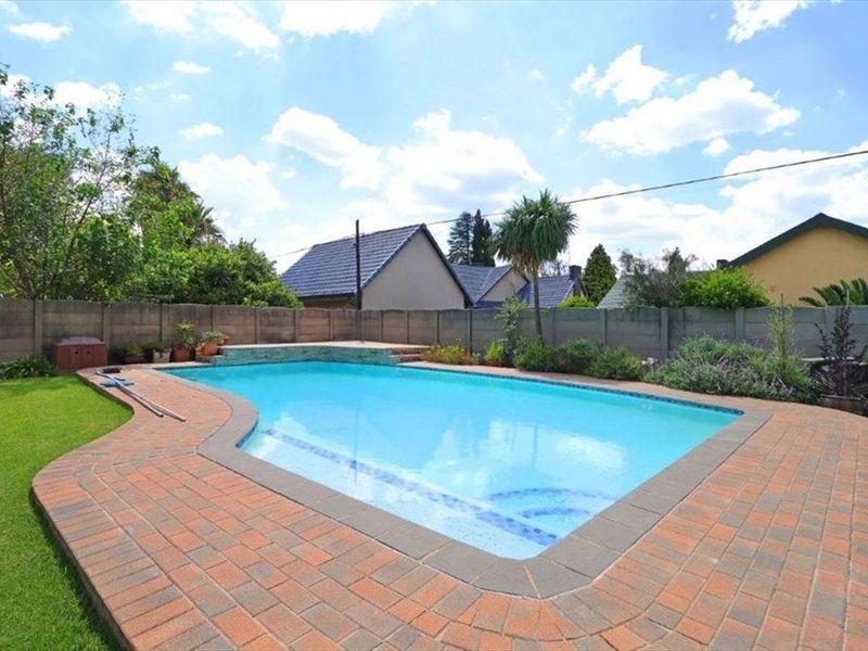 A well-maintained swimming pool area featuring brick paving surround, established garden landscaping, and a secure precast concrete boundary wall.