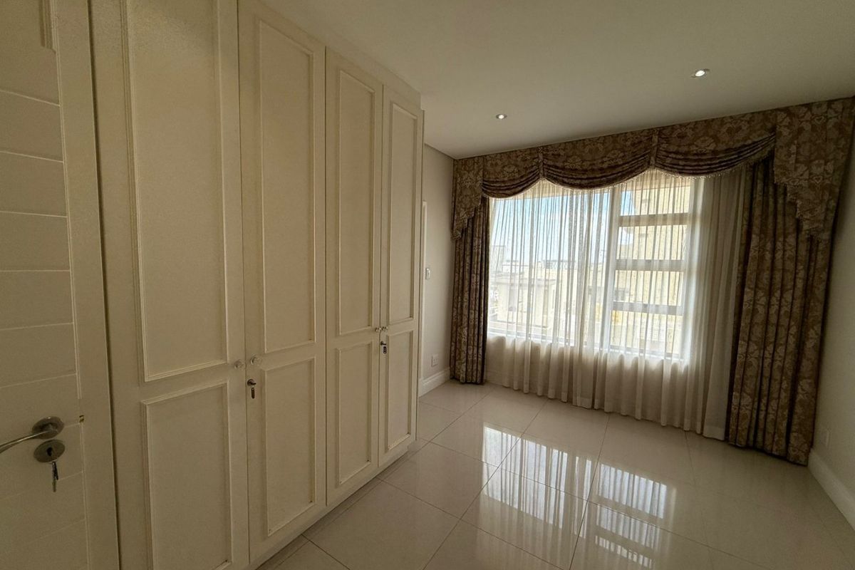 A bright and spacious bedroom featuring neutral tiled flooring, ample built-in cupboard storage, and large windows with integrated curtain pelmets.