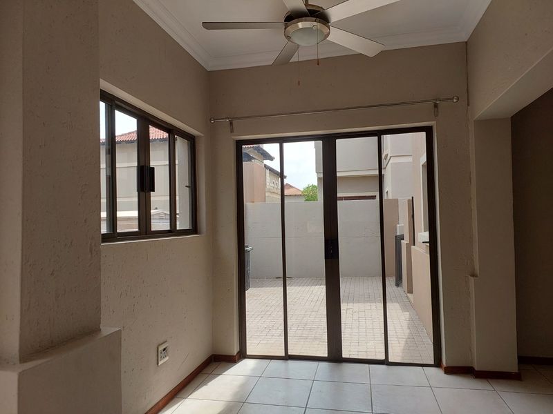 A bright, empty covered patio area featuring tiled flooring, a ceiling fan, and sliding glass doors leading to an outdoor paved courtyard.