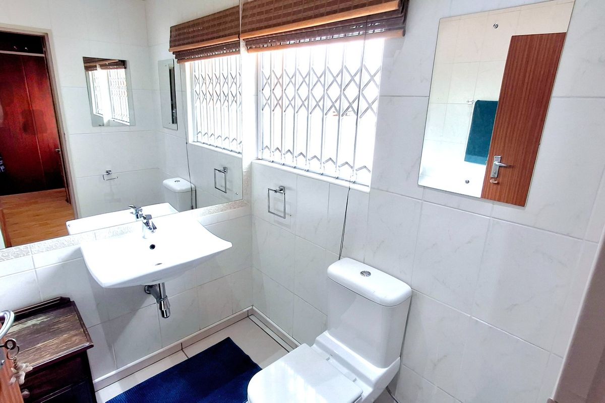 A clean, functional bathroom featuring a wall-mounted basin, toilet, and tiled finishes with security bars on the window.