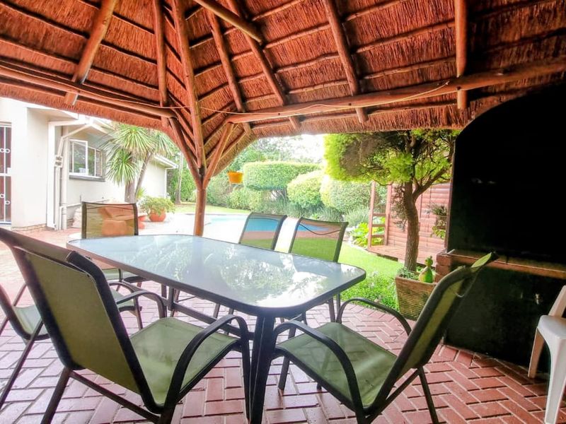 Covered outdoor patio area featuring a traditional thatched roof, brick paving, and a built-in braai, overlooking the garden and pool area.