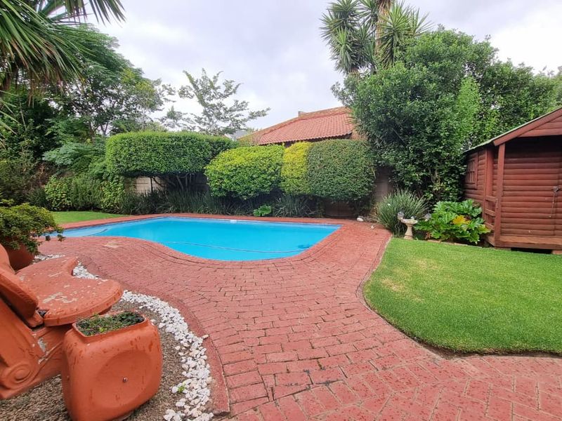 A well-maintained outdoor pool area featuring brick paving, a lush green lawn, and a wooden garden shed set within a landscaped garden.