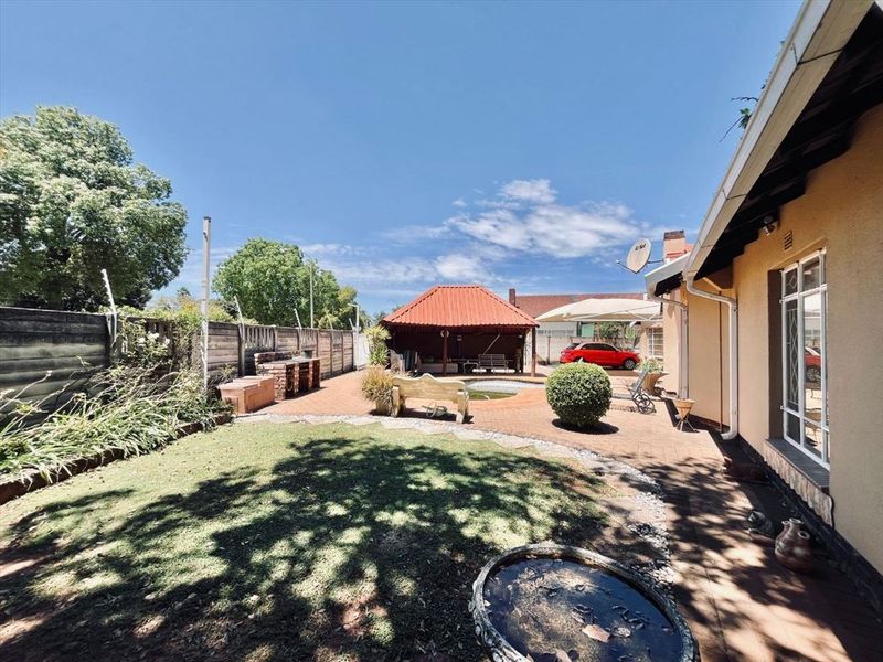 Spacious rear garden featuring a paved patio area, built-in braai, swimming pool, and a thatched gazebo, ideal for outdoor entertaining.