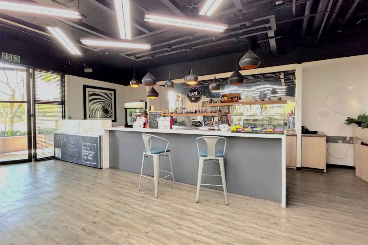 A modern commercial cafe space featuring a built-in service counter, contemporary pendant lighting, exposed ceiling, and large glass windows providing natural light.