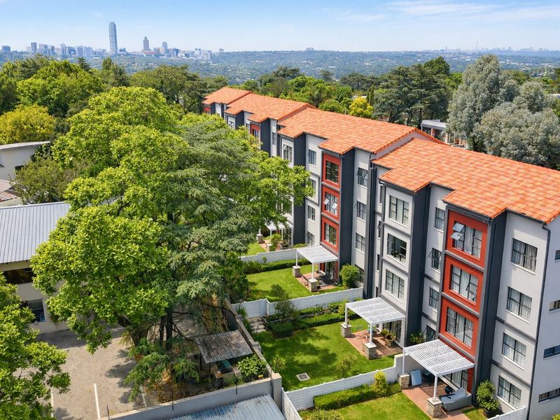 Aerial view of a modern multi-story apartment complex featuring terracotta roofing, landscaped gardens, private pergolas, and a paved parking area with city skyline views.