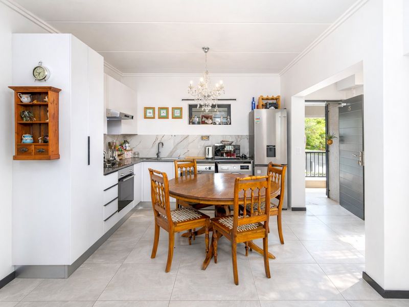 A bright open-plan kitchen and dining area featuring modern white cabinetry, stone-look backsplash, and tiled flooring, leading directly to an outdoor balcony entrance.