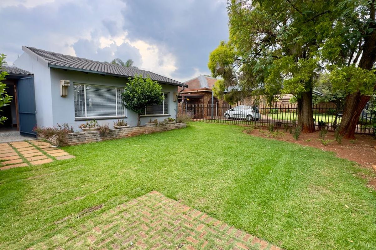 A well-maintained rear garden featuring a paved patio, a brick planter box, and established trees, enclosed by secure metal perimeter fencing.