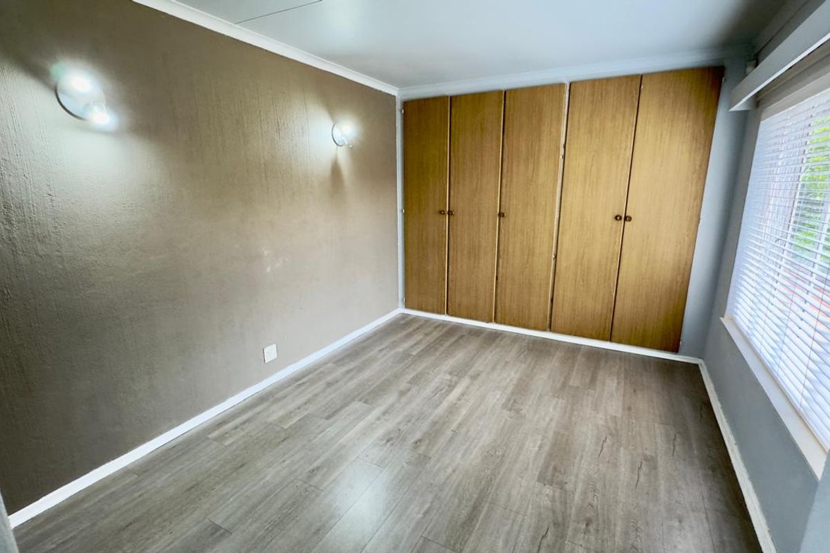 A bright, empty bedroom featuring modern laminate flooring, built-in wooden cupboards, and venetian blinds.