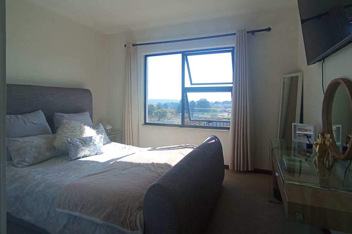 A well-lit bedroom featuring neutral carpeting, a large window with natural light, and wall-mounted television.