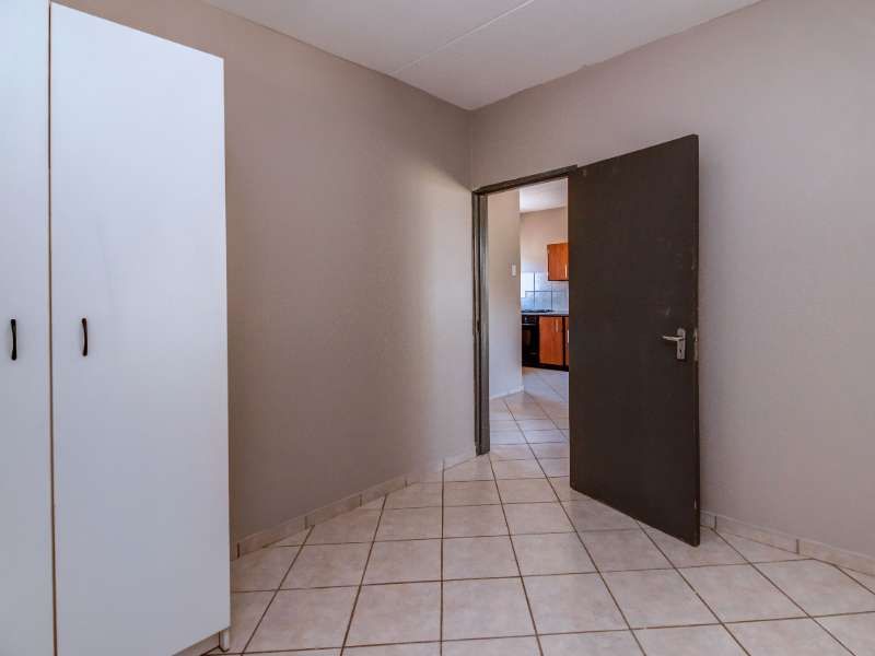 A clean, empty hallway featuring neutral wall tones, tiled flooring, and built-in storage cupboards, providing access to the adjacent kitchen area.