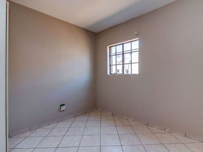 A clean, empty bedroom featuring neutral wall paint, tiled flooring, and a window fitted with security burglar bars.