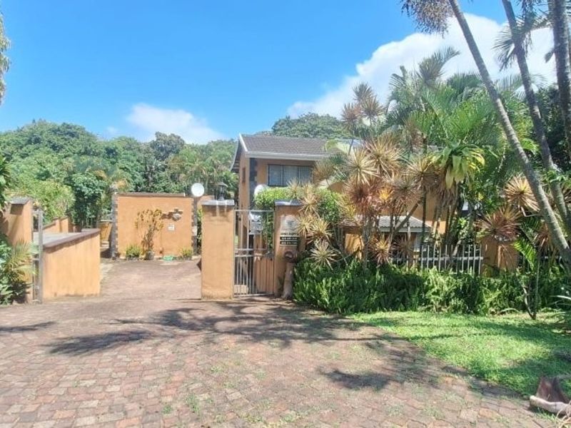 Front view of the property featuring a paved driveway, secure metal gate, and established garden surroundings.