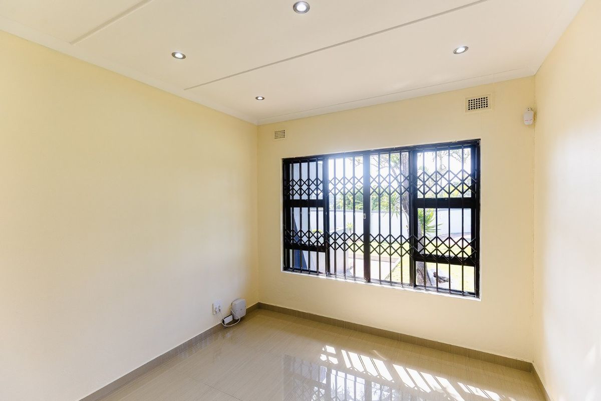 A bright, empty bedroom featuring tiled flooring, recessed downlights, and a window fitted with security bars and a gate.